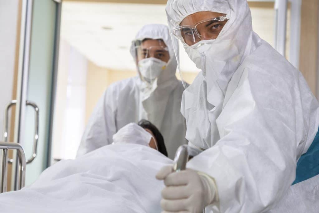 asian male nurse and doctor wearing ppe suit and face mask pushing stretcher gurney bed with seriously infected coronavirus or covid-19 patient towards quarantine room in hospital.