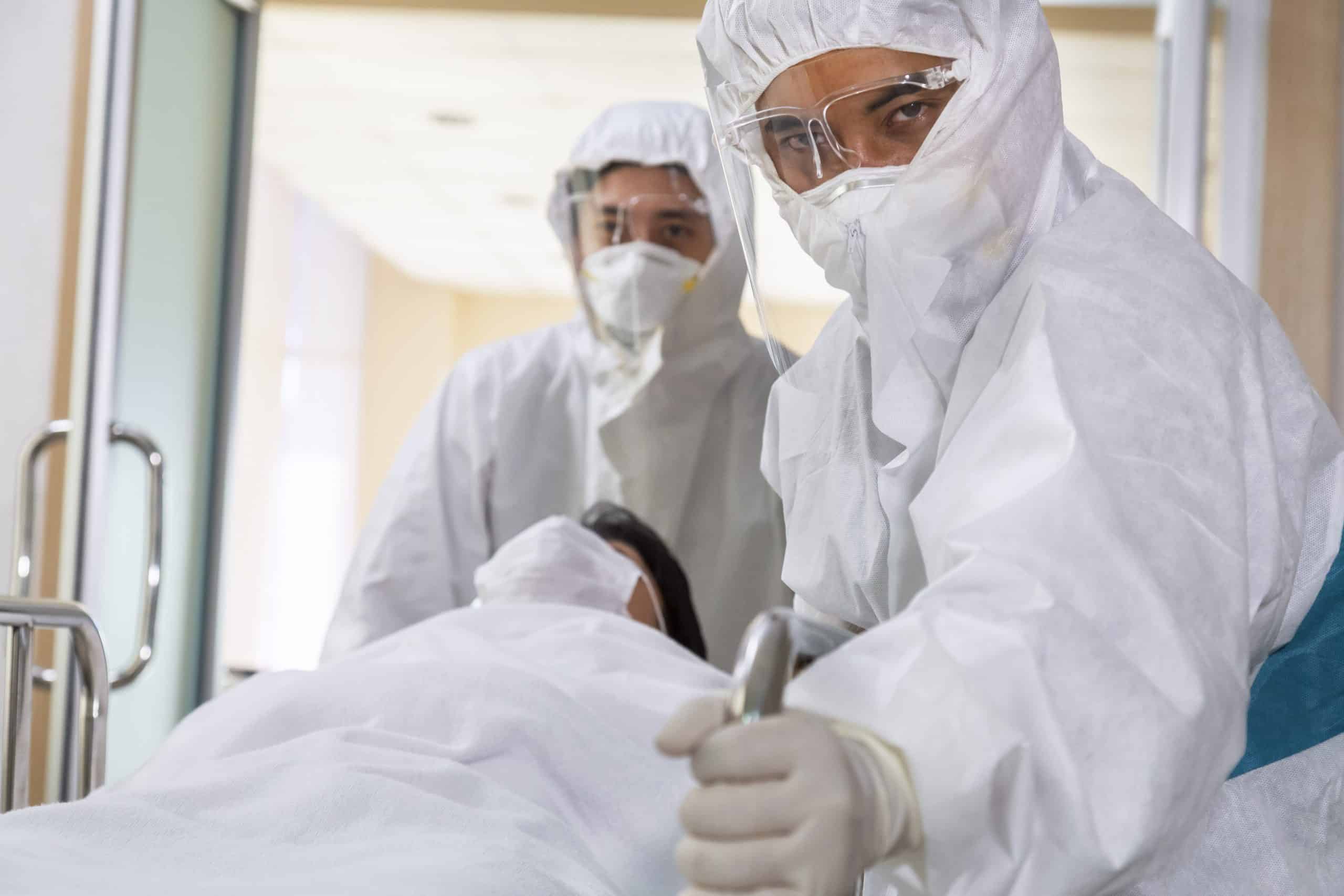 asian male nurse and doctor wearing ppe suit and face mask pushing stretcher gurney bed with seriously infected coronavirus or covid-19 patient towards quarantine room in hospital.